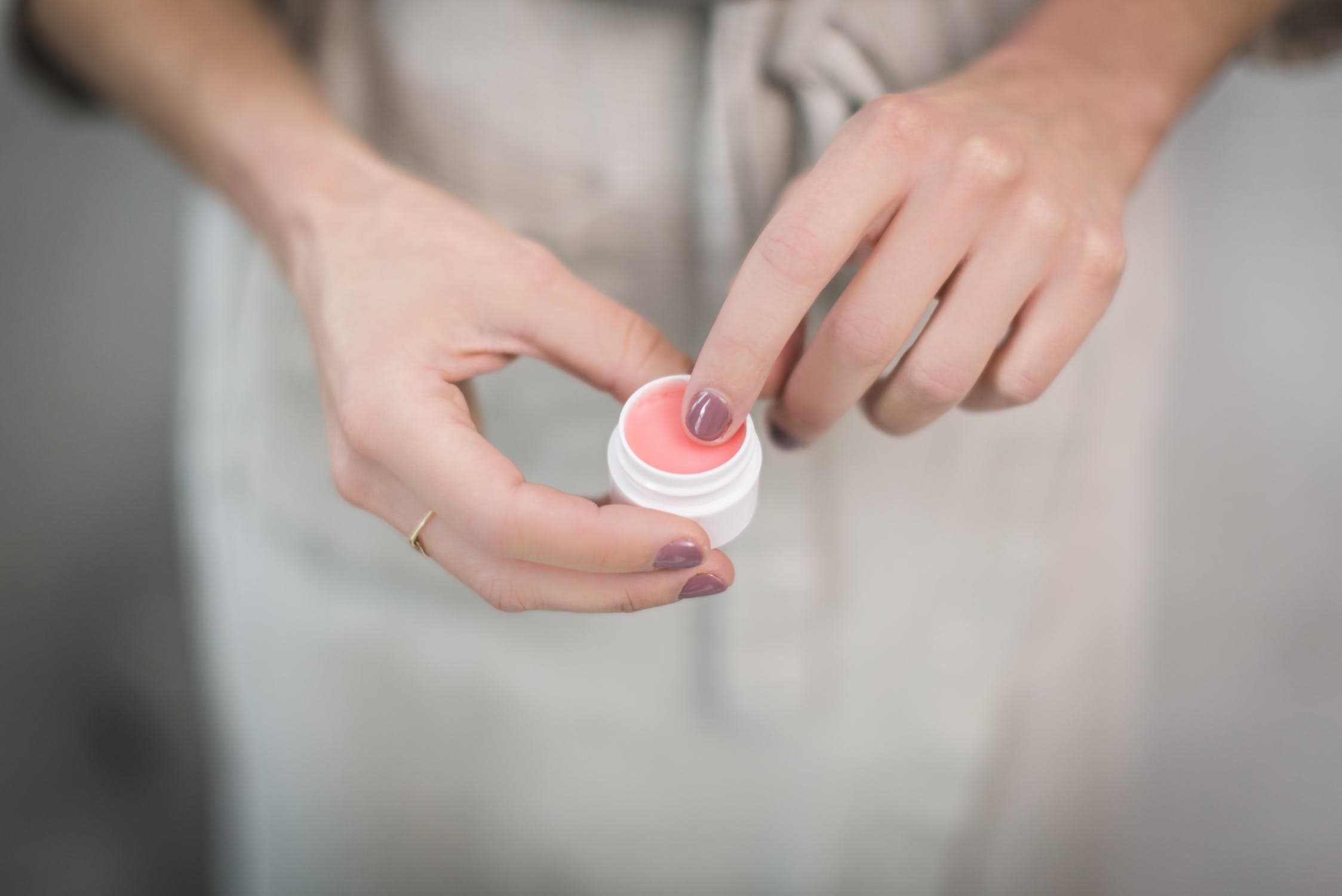 A imagem mostra uma mão feminina em destaque, segurando um pequeno pote de creme, com o dedo indicador dentro do pote