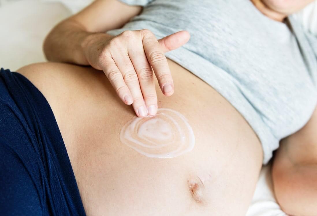 mulher grávida deitada na cama passando creme na barriga