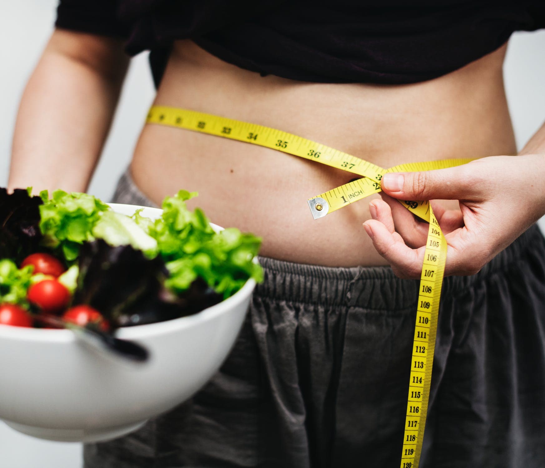 Mulher com barriga à mostra, segurando um pote de salada e com fita métrica medindo sua cintura.
