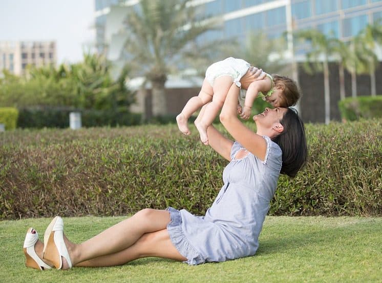 A imagem mostra uma mulher, sentada no gramado em ambiente externo, segurando o filho suspenso e sorrindo