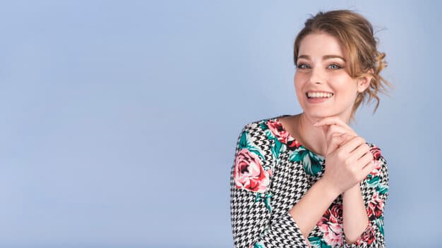 mulher sorrindo com as mãos embaixo do queixo com uma camisa florida em frente a um fundo azul