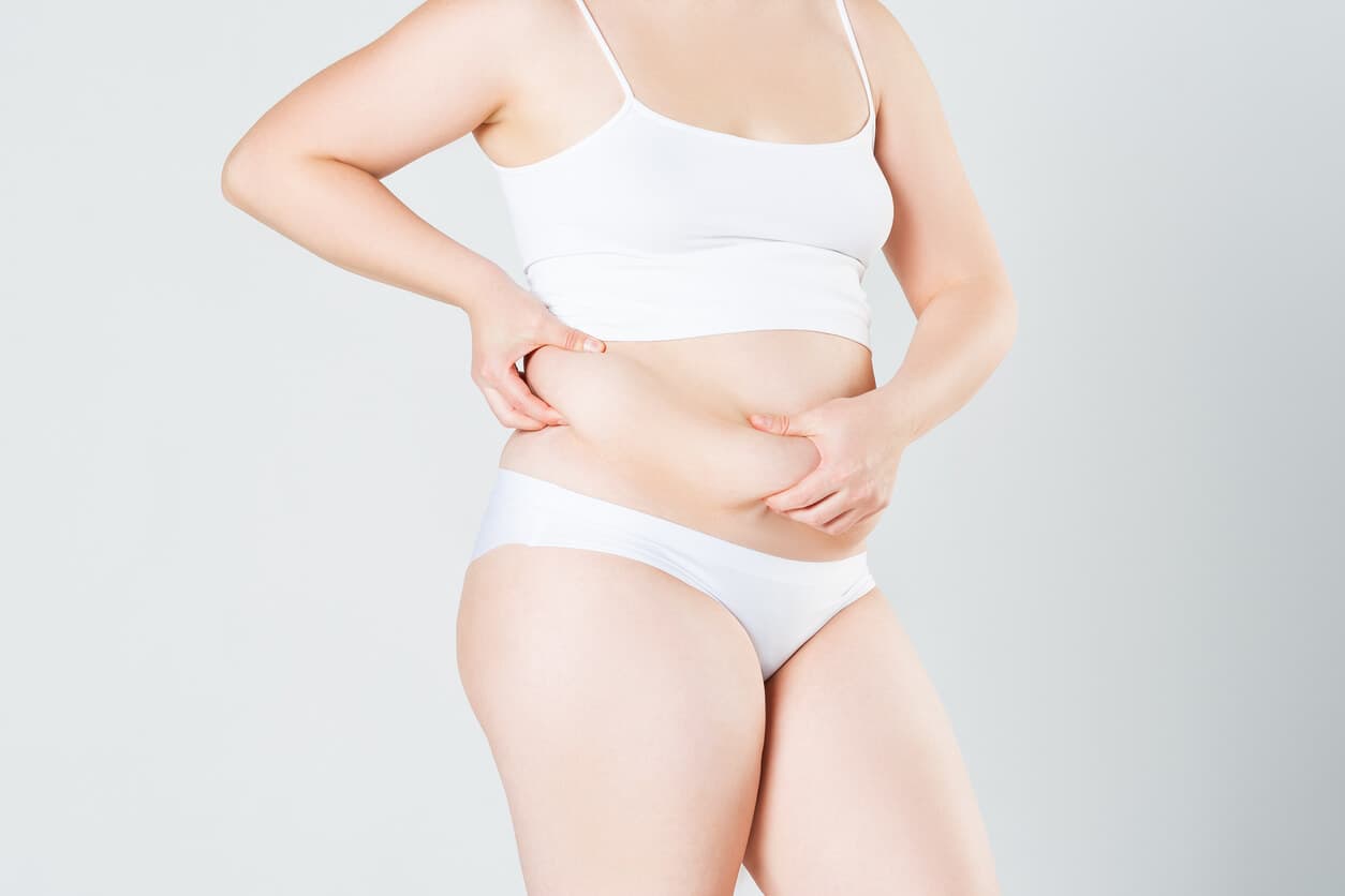 Mulher de roupas íntimas brancas segurando o excesso de pele e gordura na barriga.