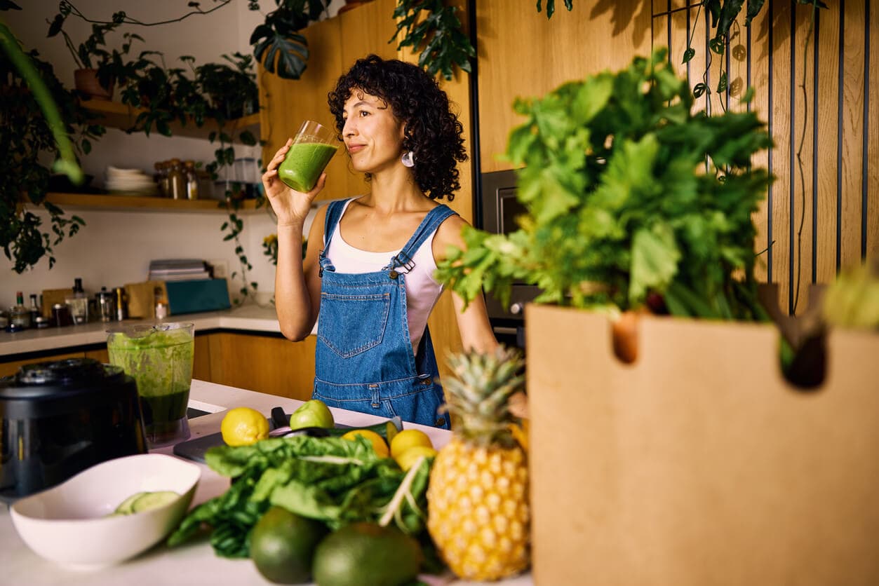 Mulher tomando suco verde em uma cozinha com frutas, verduras e folhas frescas sobre a bancada.