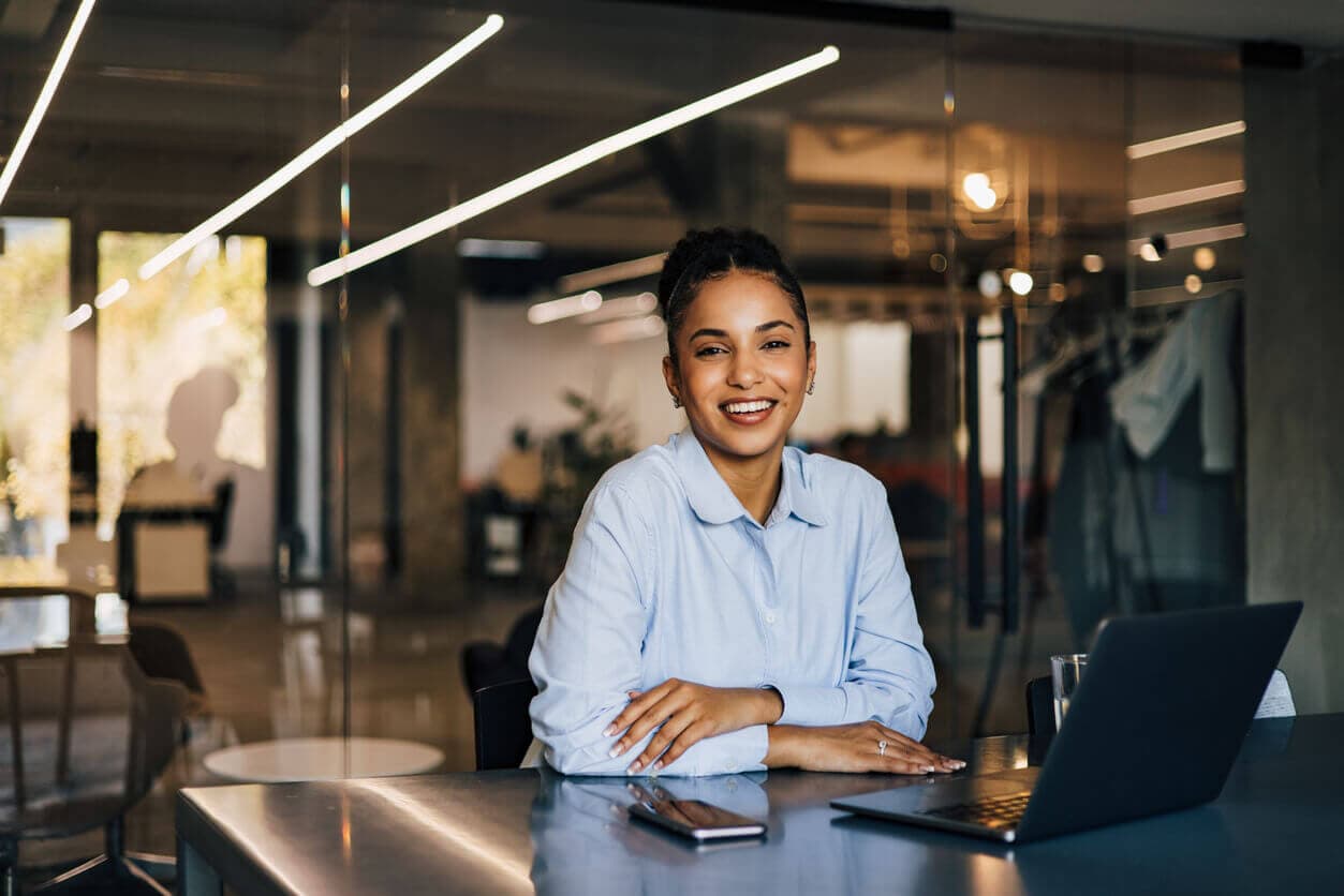 Mulher sorridente trabalhando em escritório moderno, sentada à mesa com notebook e celular.