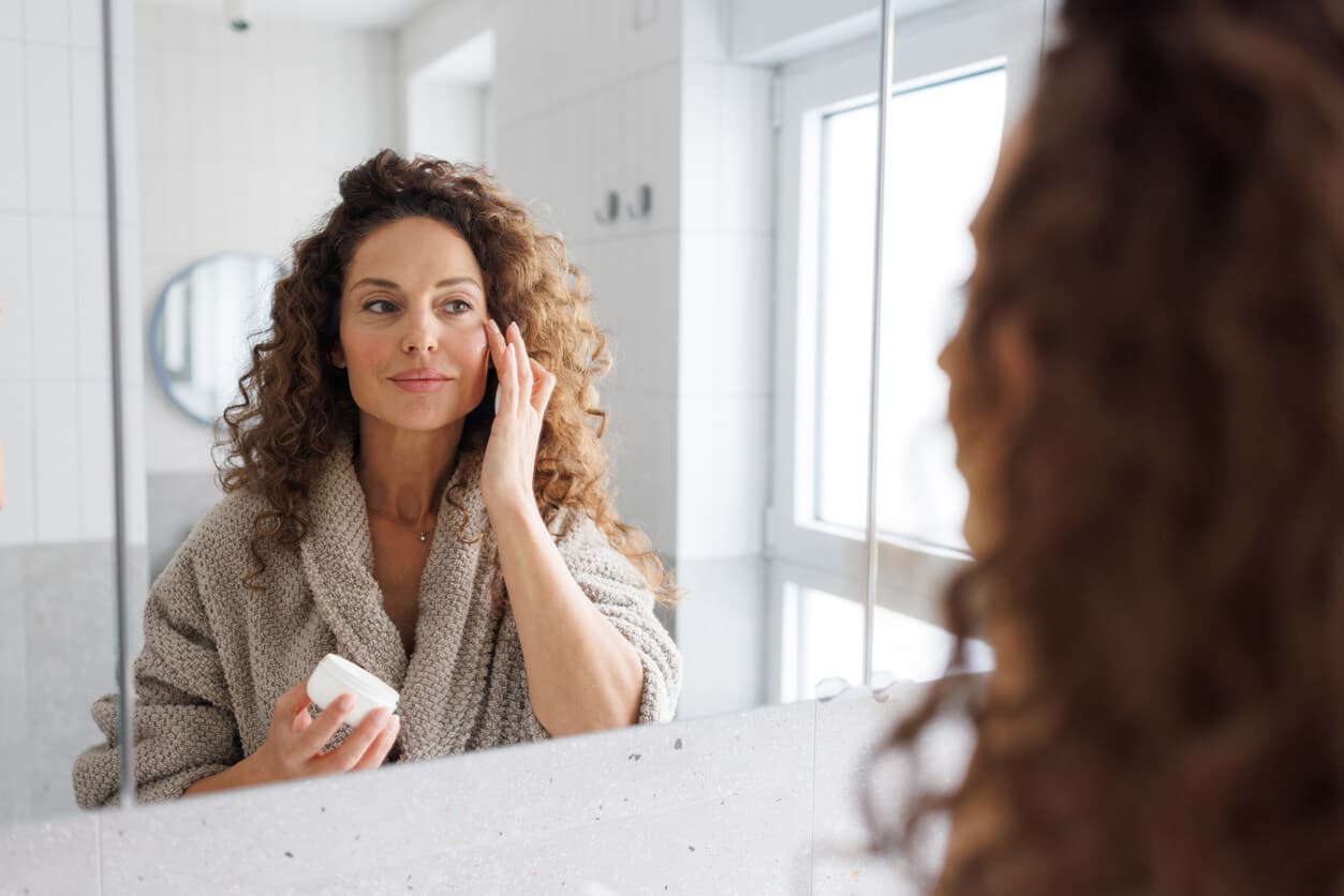 Mulher aplicando produto de skincare no rosto em frente ao espelho