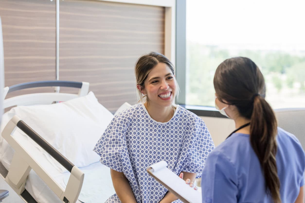 Paciente sorridente em quarto hospitalar conversa com profissional de saúde ao lado da cama durante orientação no pós-operatório.