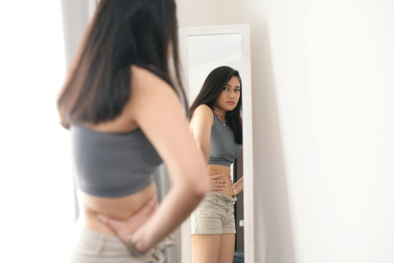 Mulher em frente ao espelho observando o próprio corpo, com expressão séria, em ambiente interno claro.
