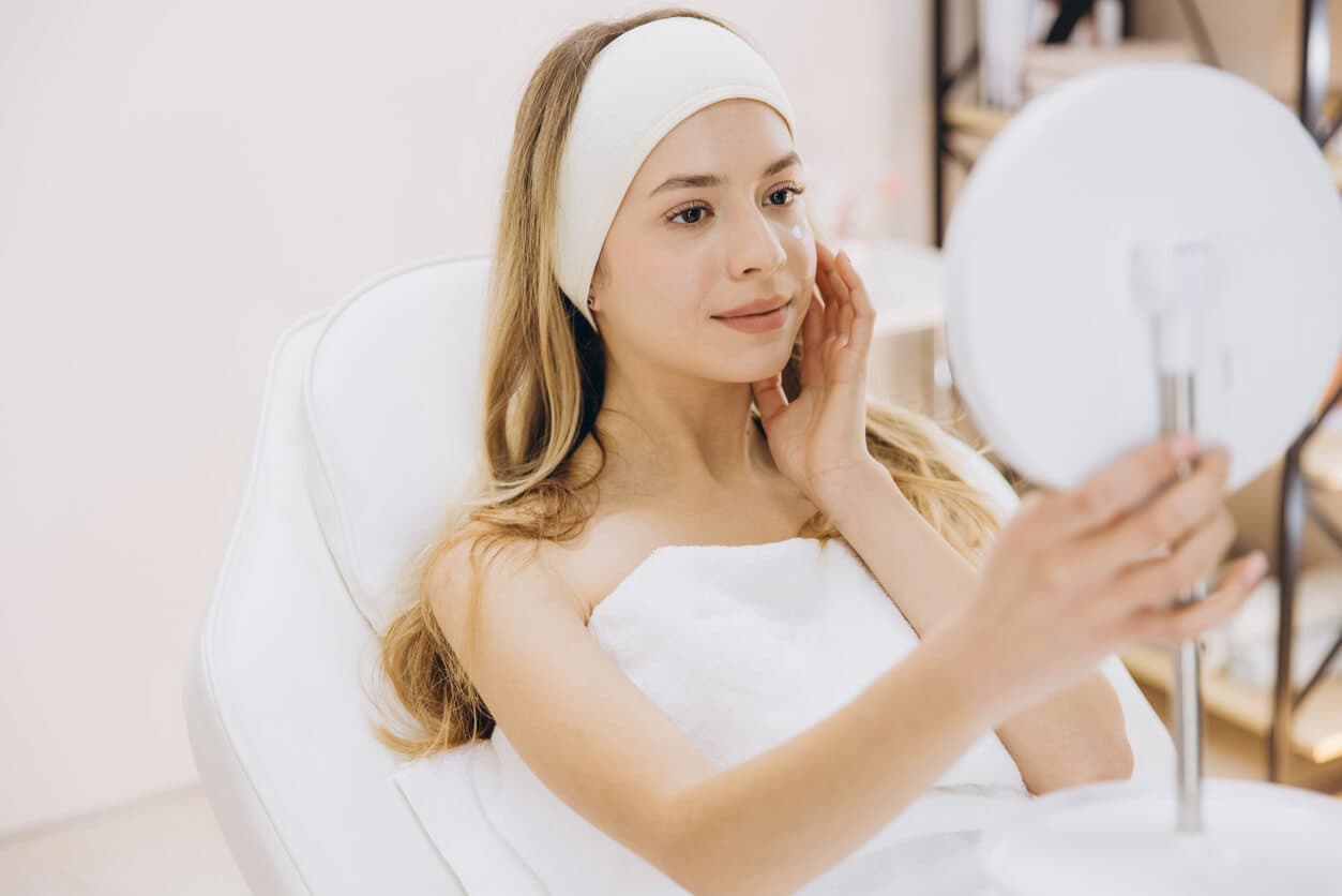 Mulher sentada em cadeira de atendimento estético, com faixa no cabelo e toalha branca, observando o rosto no espelho enquanto toca suavemente a pele da face.
