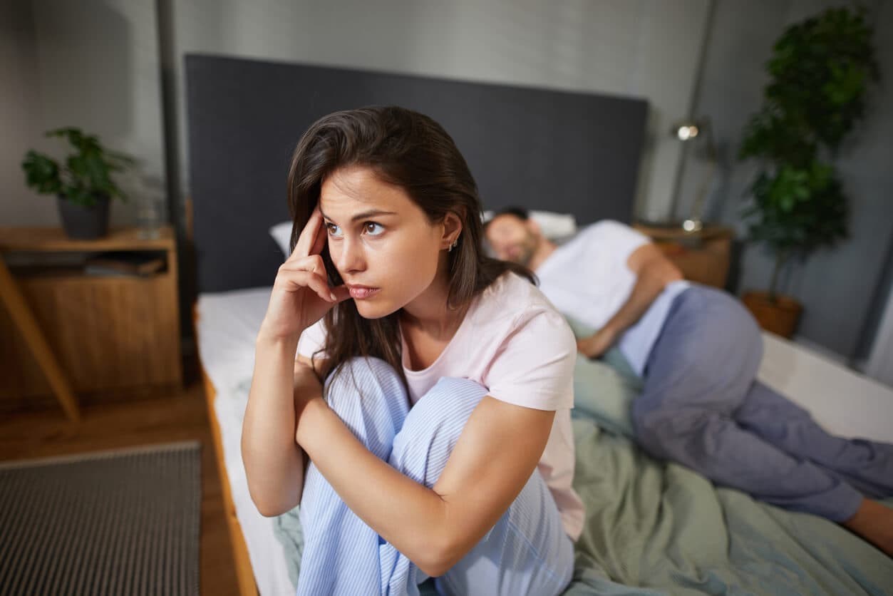 Mulher triste sentada na cama enquanto um homem dorme ao fundo