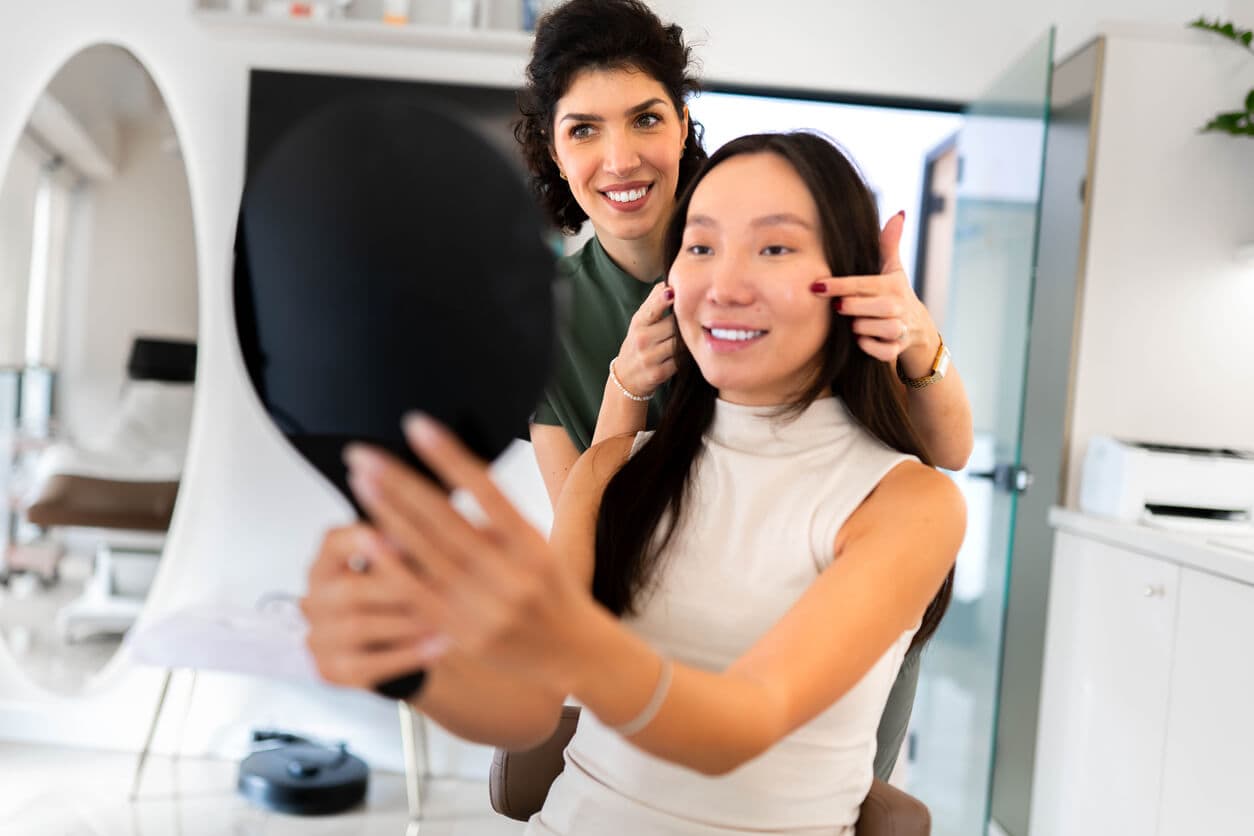 Profissional de estética mostrando os resultados de um tratamento facial sem cortes para paciente que se observa no espelho