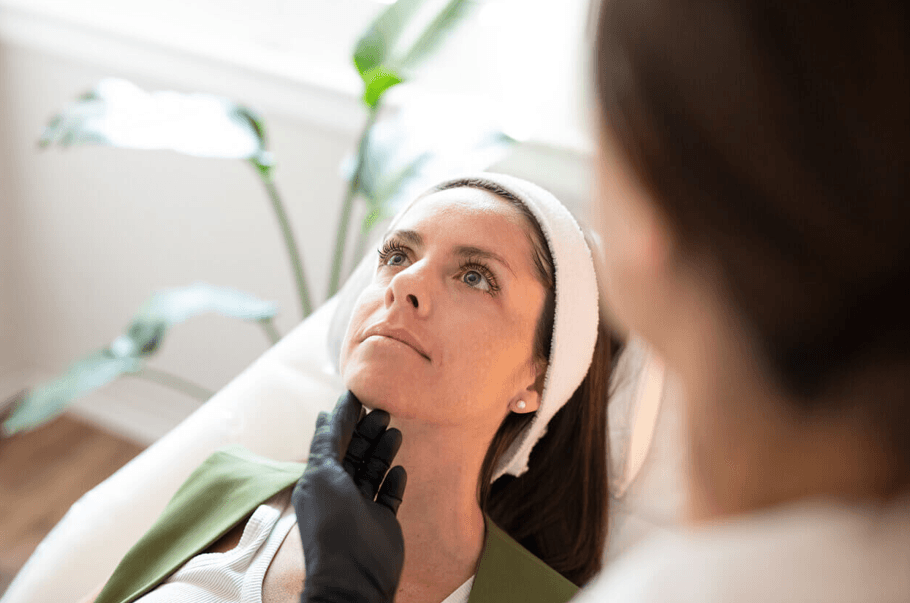 Mulher em atendimento estético facial, deitada em maca enquanto profissional avalia a região do rosto e do pescoço.