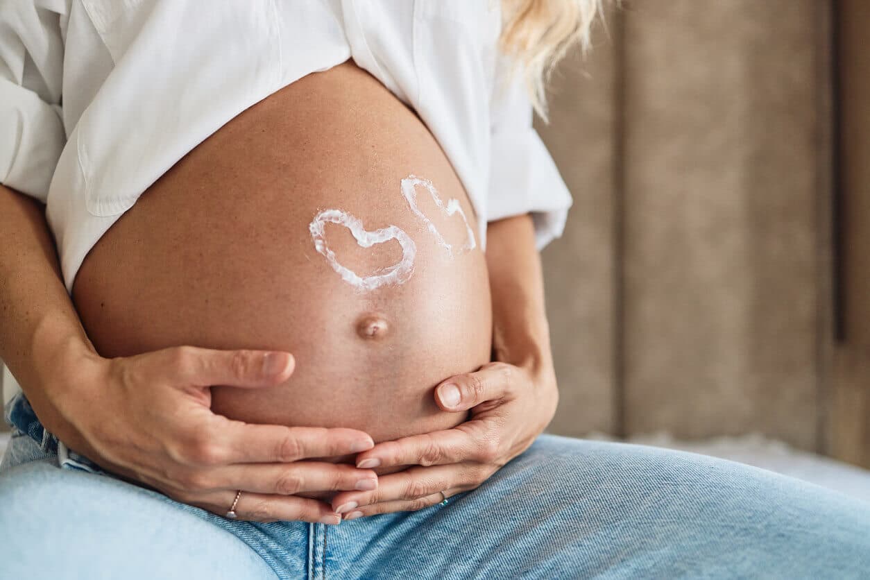 Mulher grávida sentada, com as mãos apoiadas sobre a barriga e um desenho de corações feitos com creme na pele do abdômen.