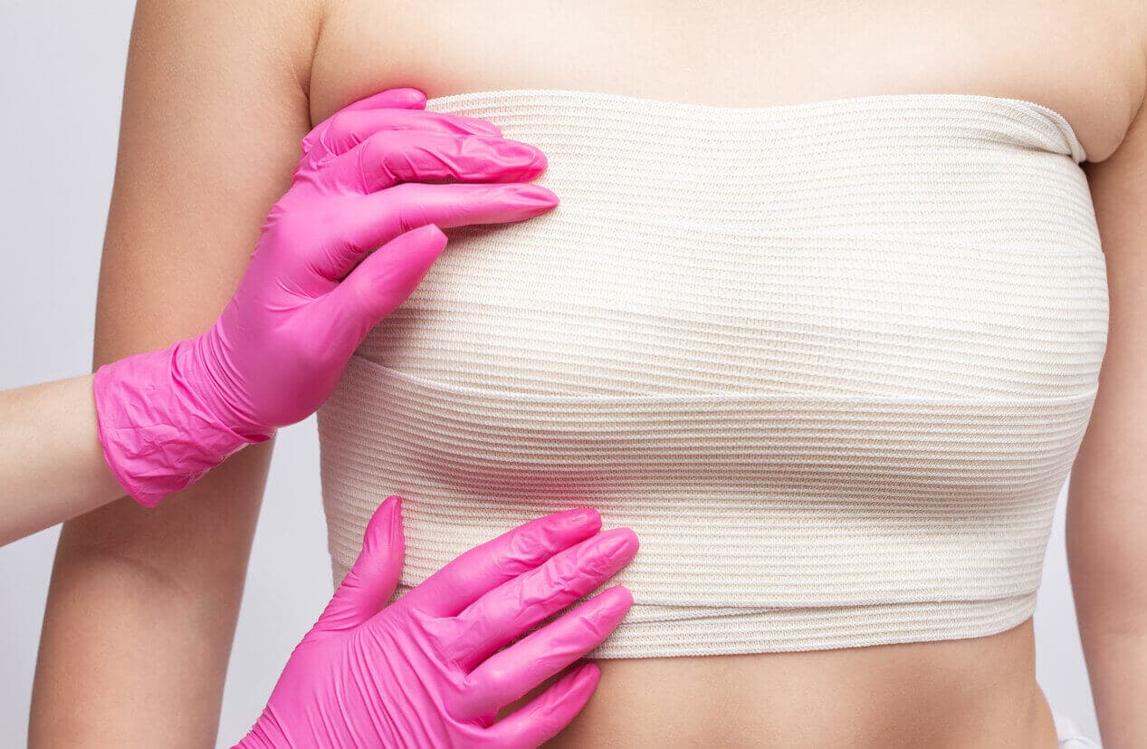 Profissional de saúde com luvas rosas avaliando região torácica de paciente com curativo compressivo, ilustrando cuidados no pós-operatório de cirurgias plásticas.
