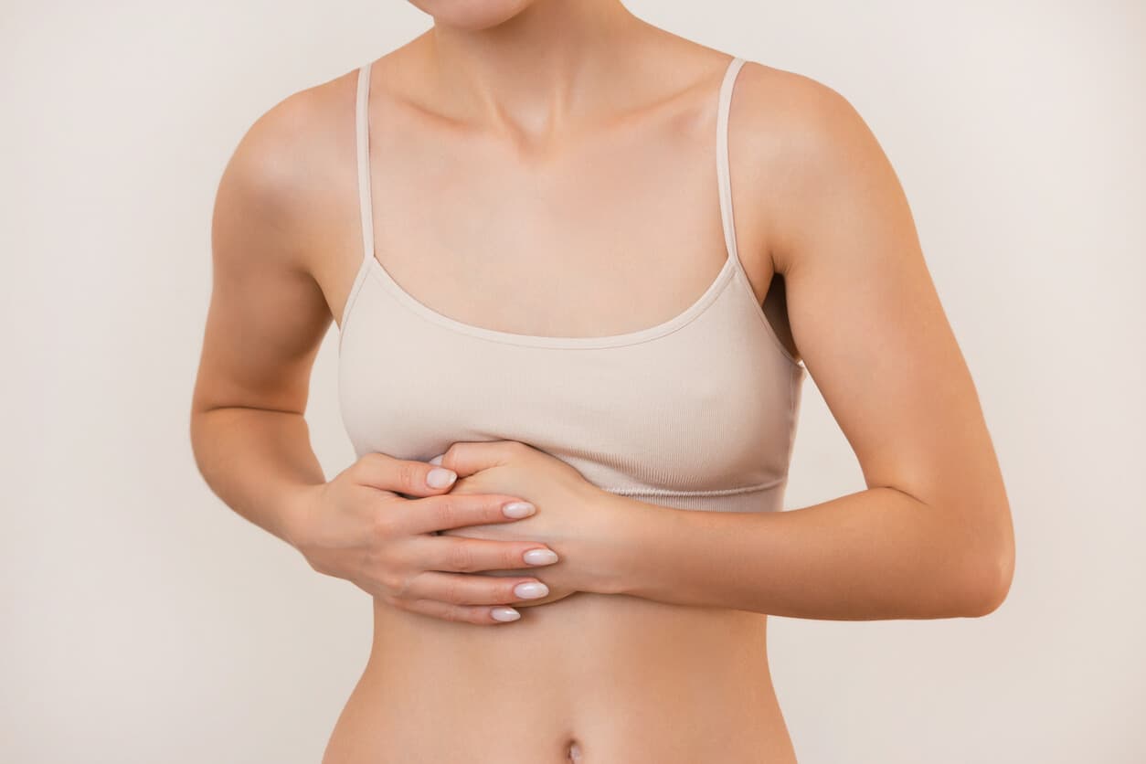 Mulher usando top bege segura a região inferior da mama com as mãos, gesto que pode indicar desconforto ou a presença de mama acessória.