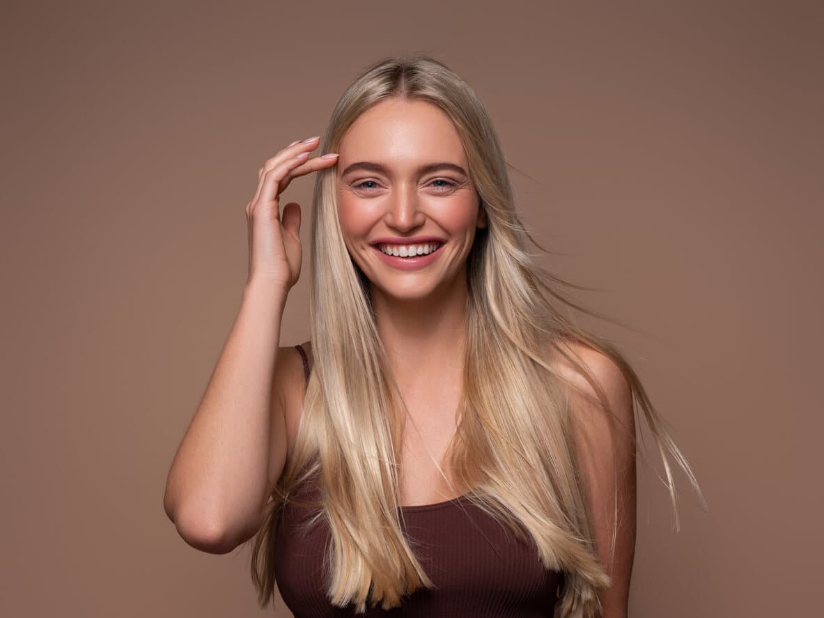 Mulher jovem e loira sorrindo e levando a mão aos cabelos