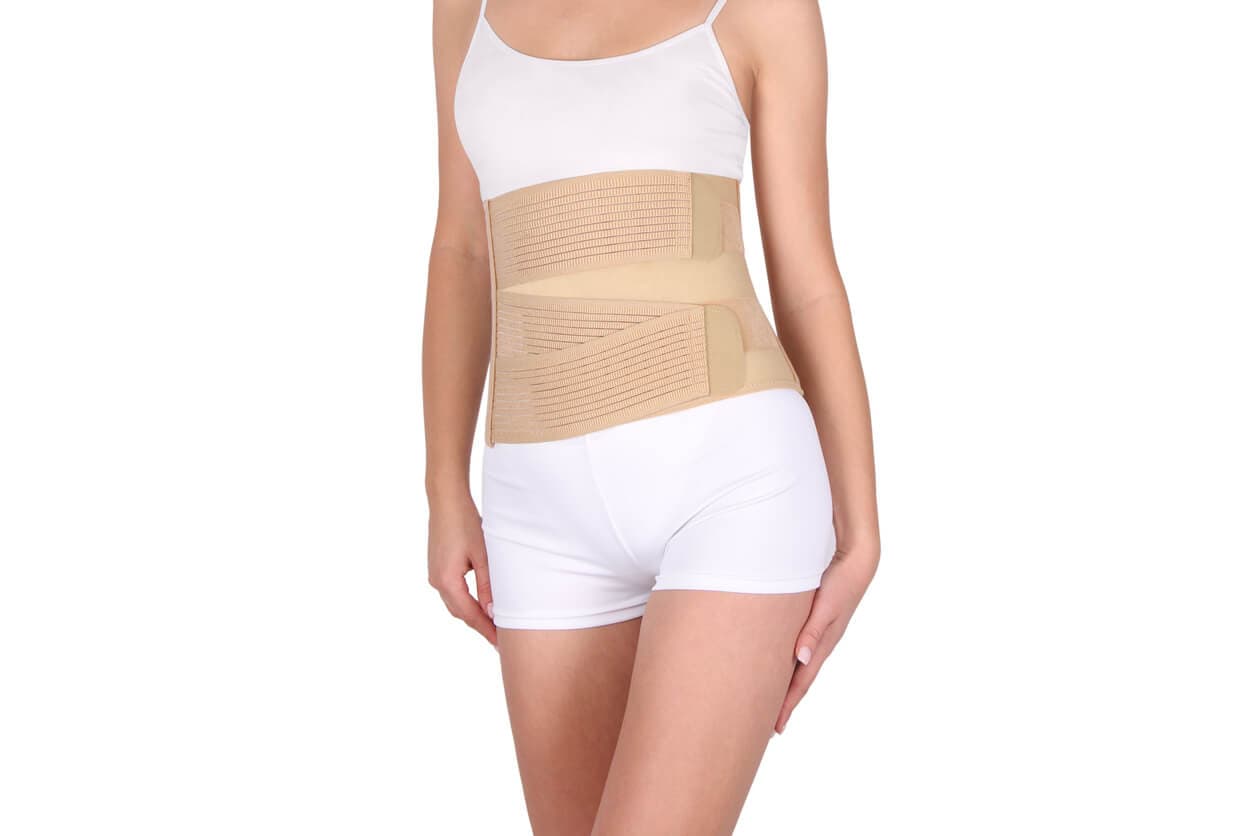 Mulher usando cinta abdominal pós-cirúrgica bege com fechamento em velcro sobre top branco e shorts brancos