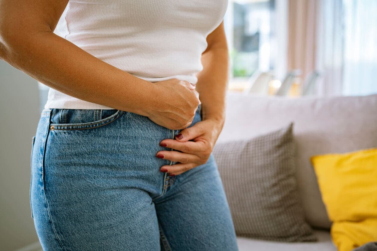 Mulher em pé, usando regata branca e calça jeans, com a mão pressionando a região íntima por desconforto