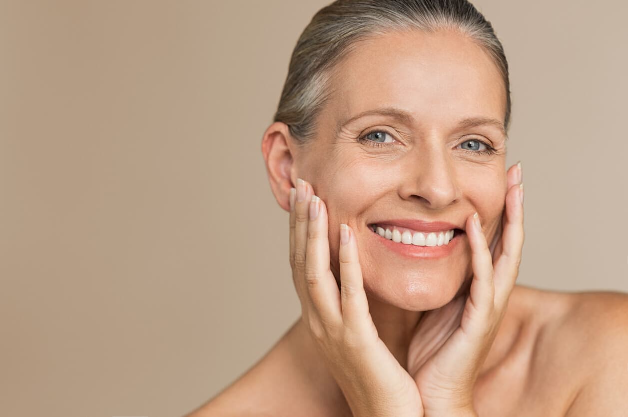 Mulher madura sorrindo, com as mãos apoiadas no rosto, em close e fundo bege, destacando a pele da face e a região mandibular.