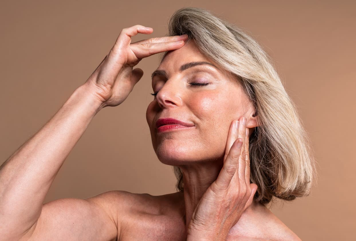 Mulher madura de olhos fechados, com as mãos na testa e na lateral do rosto, em pose que destaca a região facial e o contorno da mandíbula.
