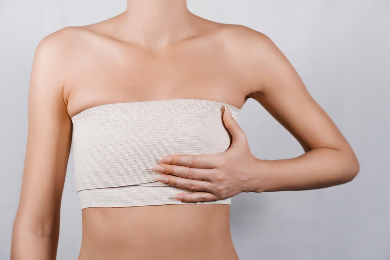 Mulher com faixa cirúrgica no tórax cobre os seios com a mão, representando recuperação após procedimentos de mamoplastia e mastopexia.