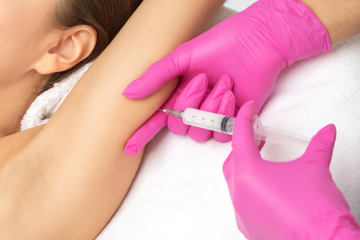 Profissional com luvas rosas aplica injeção no braço de paciente durante procedimento no braço.
