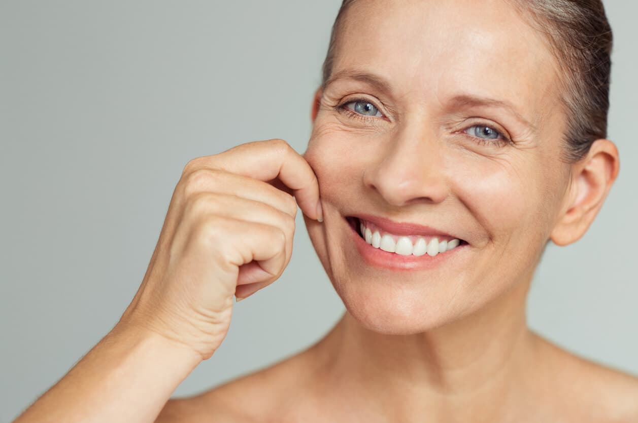 Mulher madura sorrindo enquanto segura levemente a pele da bochecha, ilustrando flacidez facial e sinais de envelhecimento no rosto.