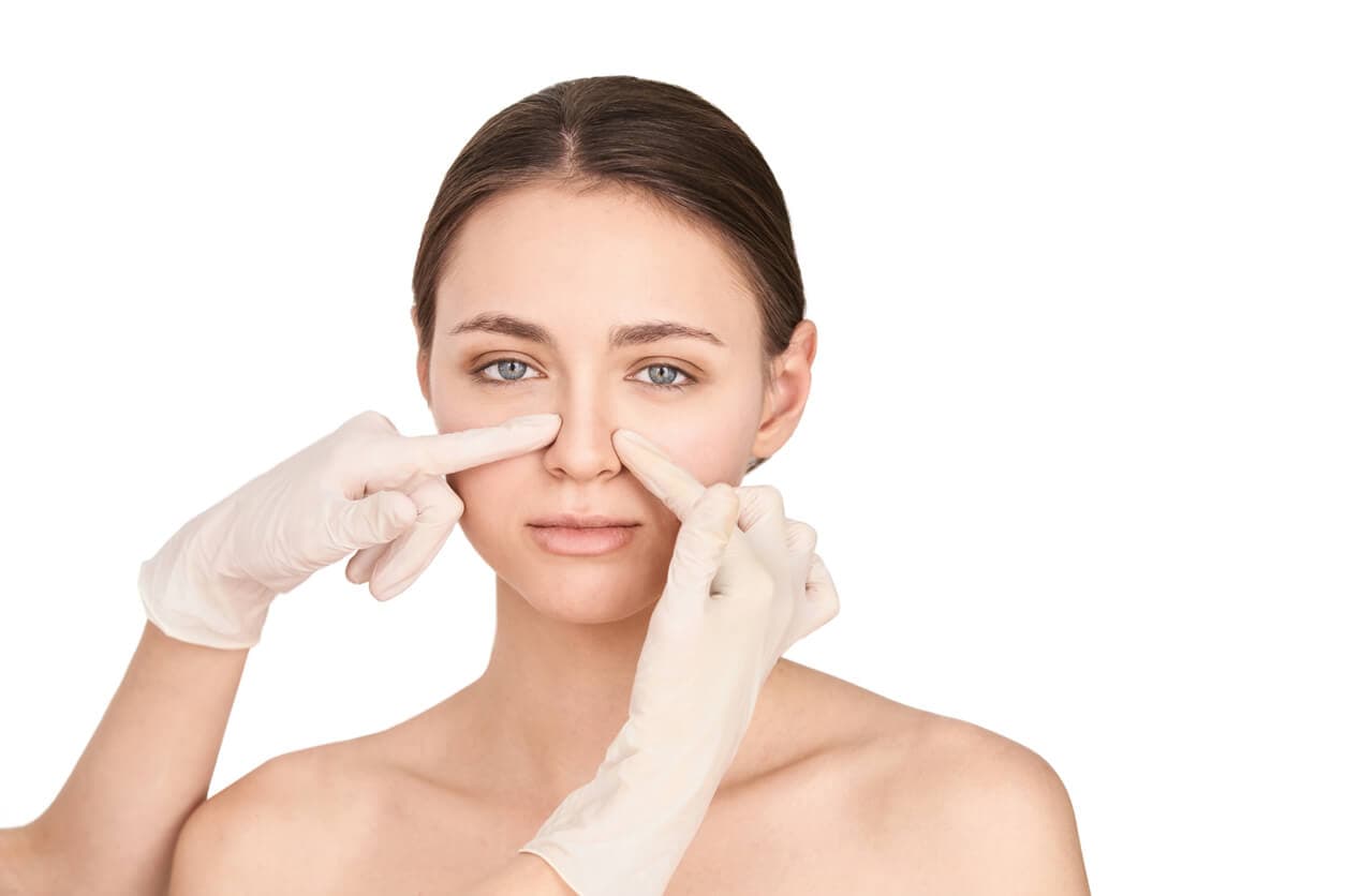 Mulher com expressão neutra, enquanto duas mãos com luvas tocam lateralmente o nariz, sugerindo avaliação estética da região nasal.