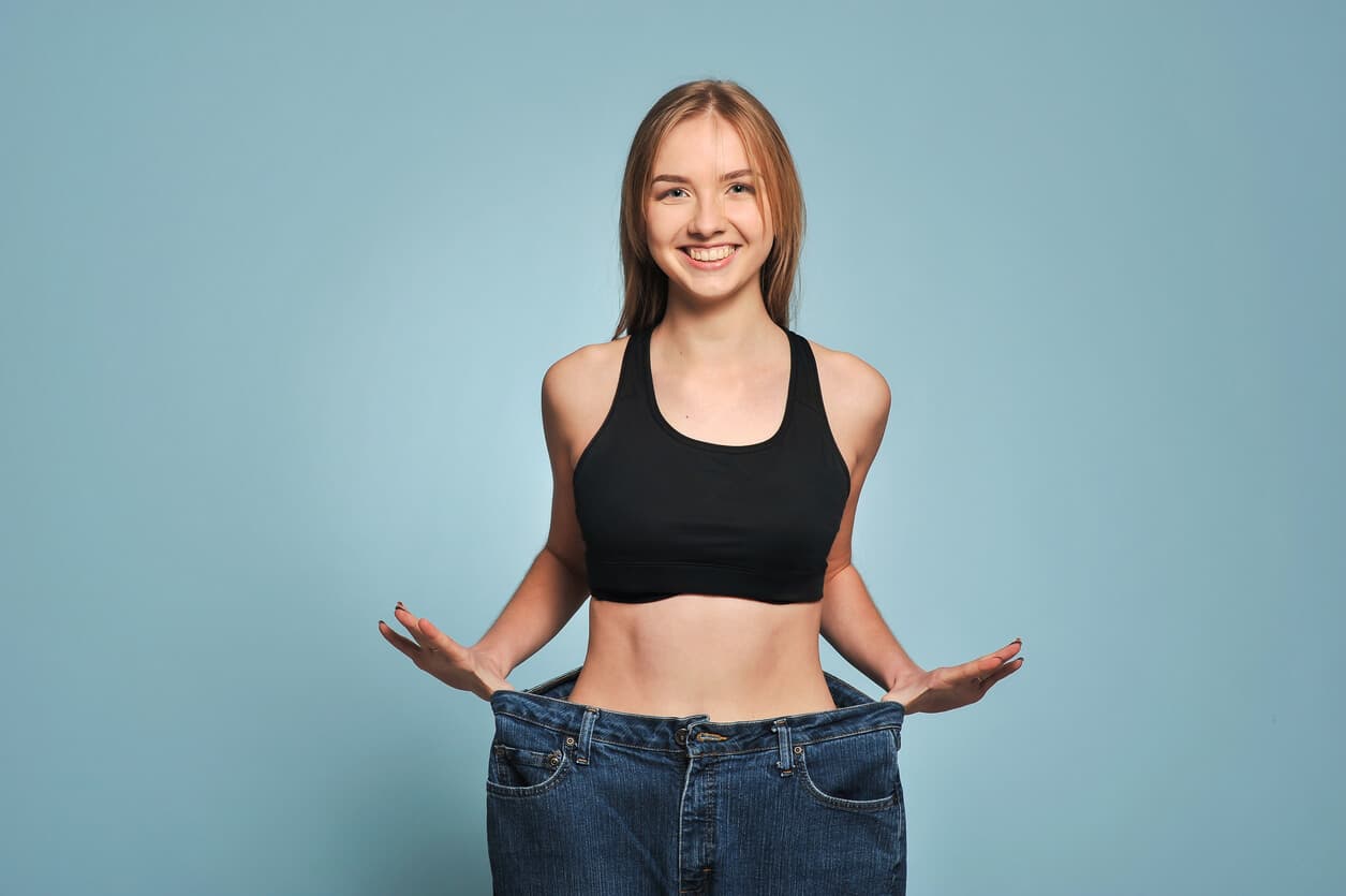 Mulher jovem com top esportivo segura calça jeans larga, destacando redução de medidas e contorno abdominal mais definido.