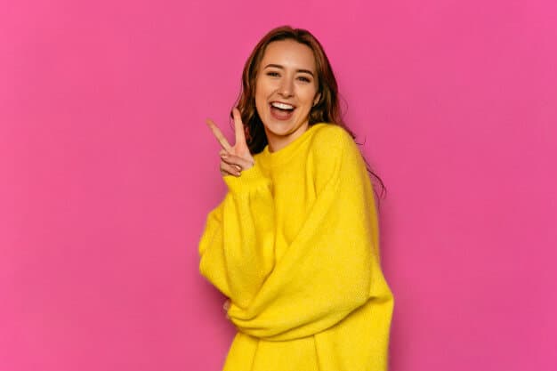 menina fazendo sinal de paz sorrindo de blusa amarela em frente a um fundo rosa